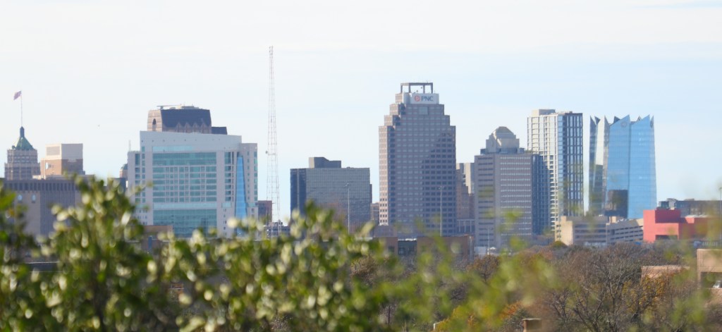 Daytime photograph of the downtown San Antonio skyline, with buildings and trees in the foreground
